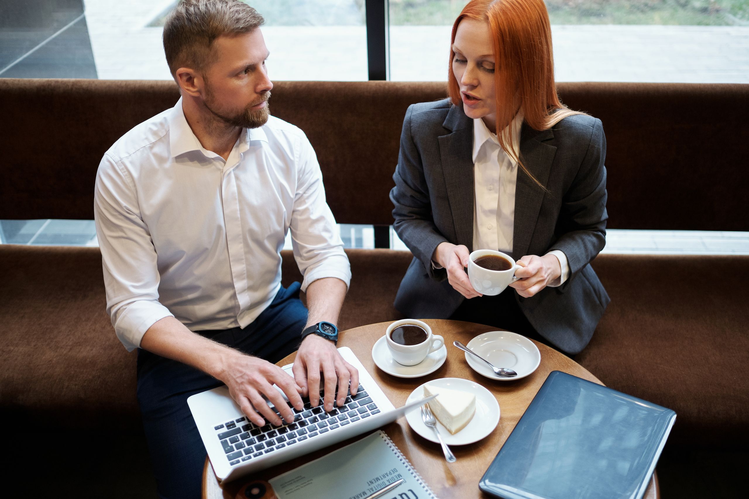 A man and a woman talk over coffee while he types on a laptop