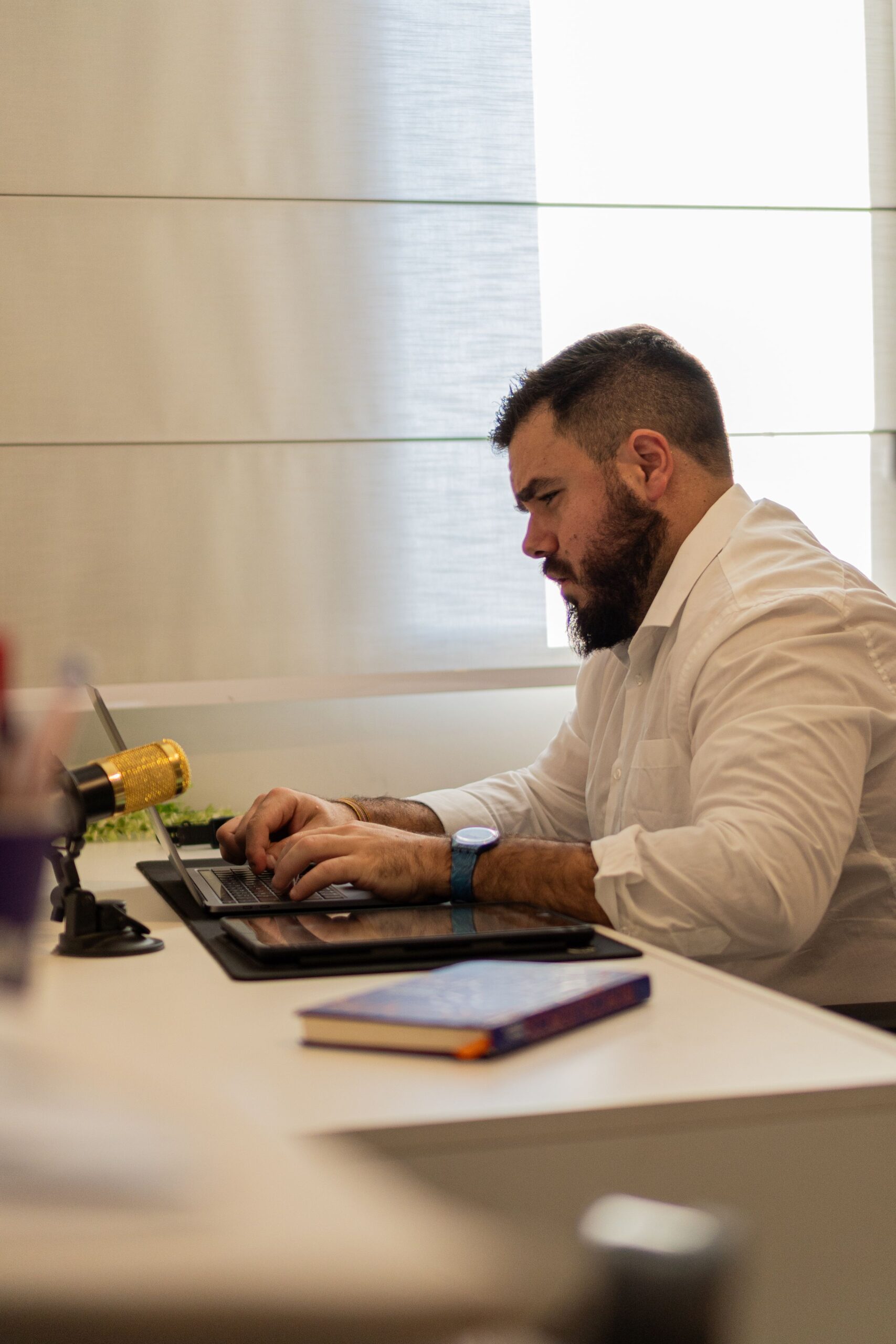 A man in a white shirt works at a laptop beside a bright window