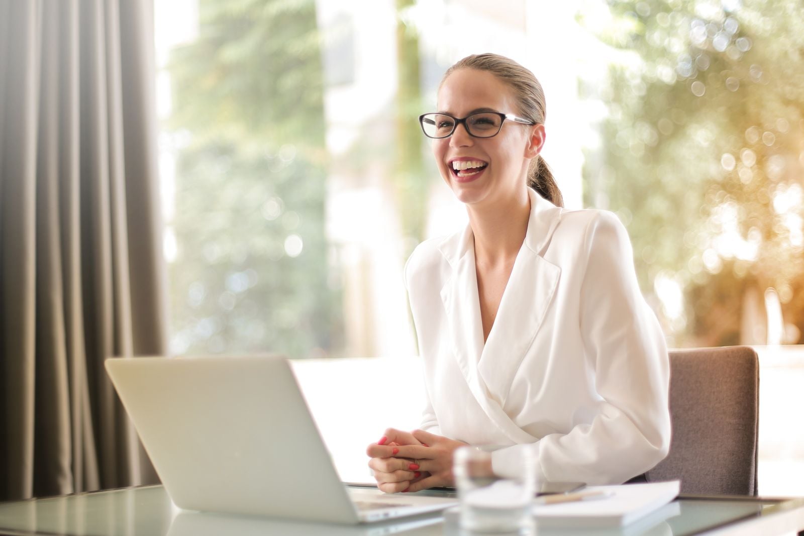 A business owner laughing at her laptop beside a bright window with greenery outside
