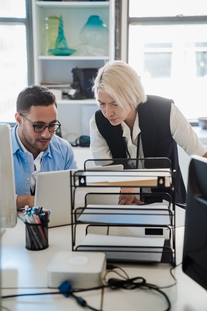 Two colleagues at a desk looking at a laptop together, one leaning in to check the screen