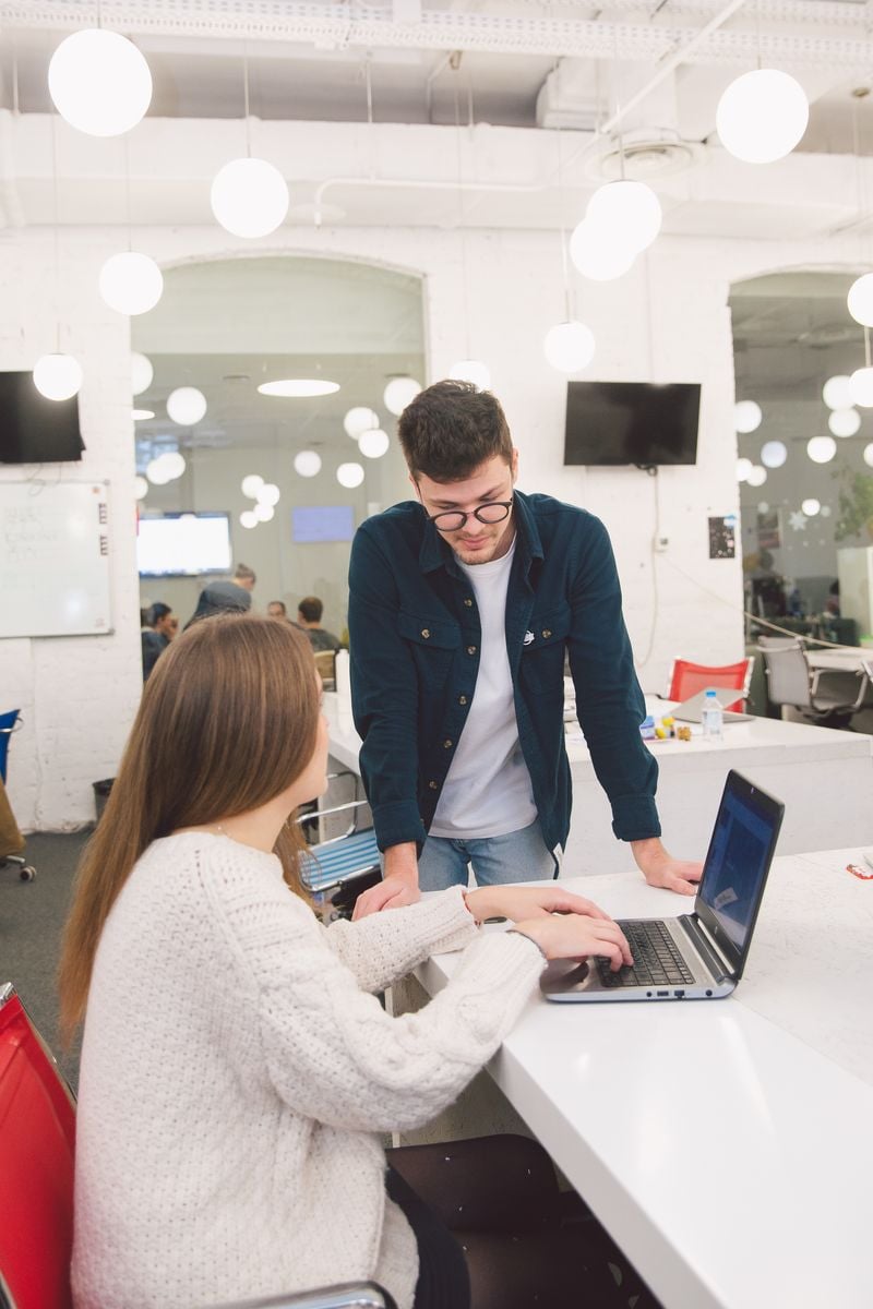 A man stands beside a seated colleague, looking at her laptop together in a bright open office