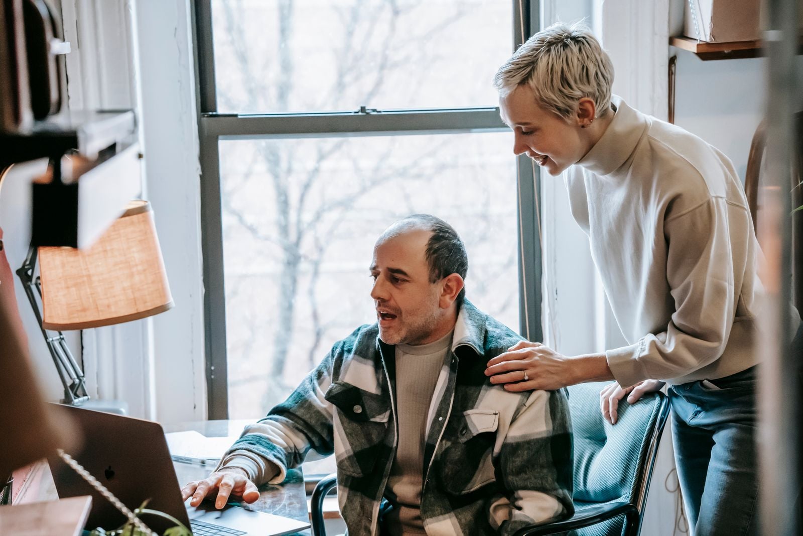 A woman leans over a colleague's shoulder, smiling as she helps him with his laptop by a bright window