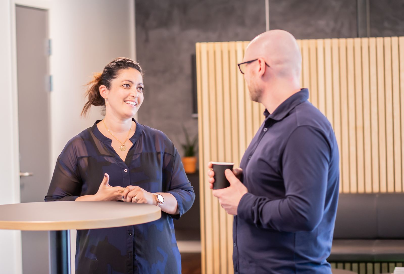 A smiling woman chats with a colleague who holds a coffee, standing at a small table in a bright office